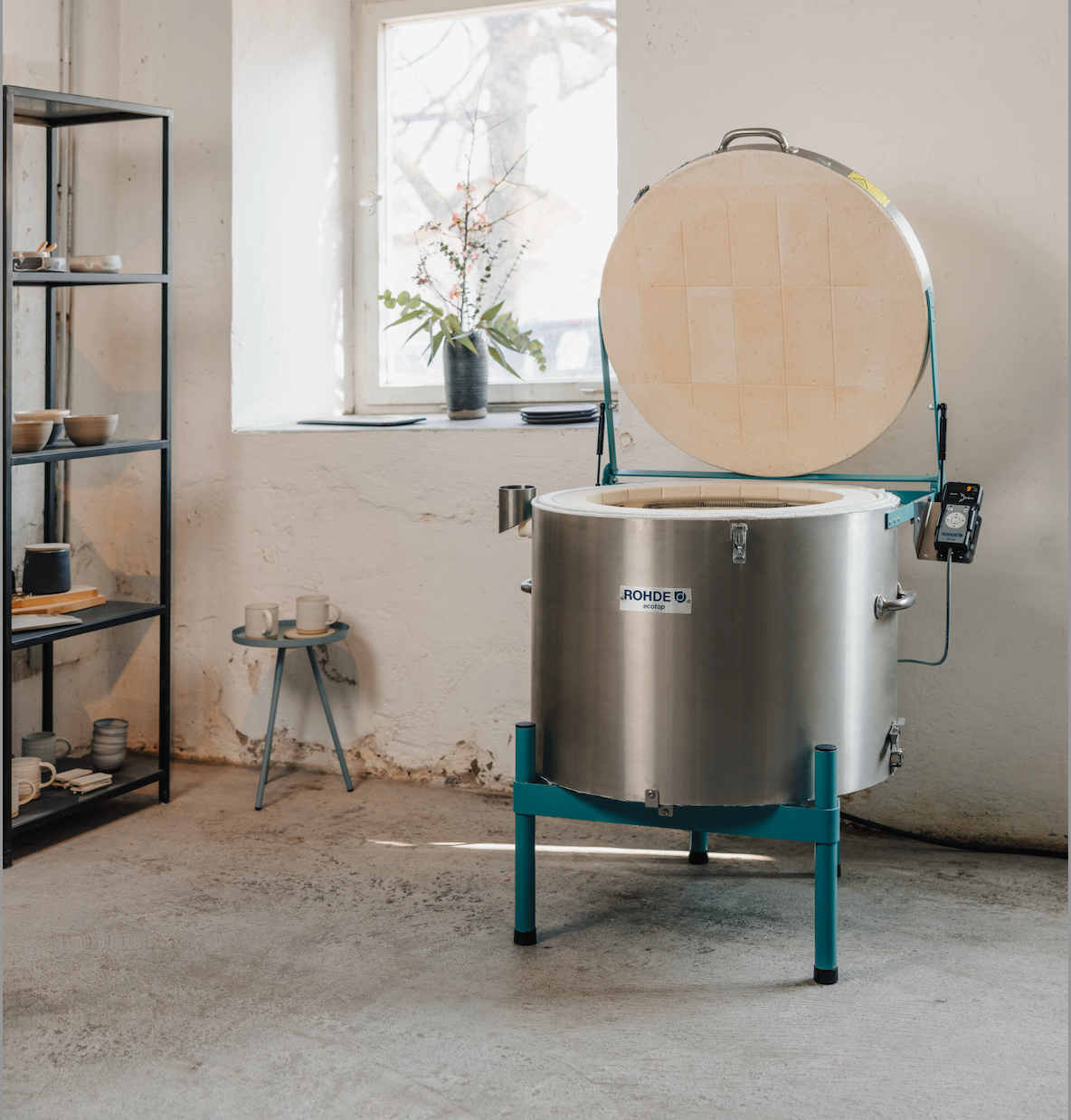 Stainless steel kiln with open lid in a room with shelves and a window.